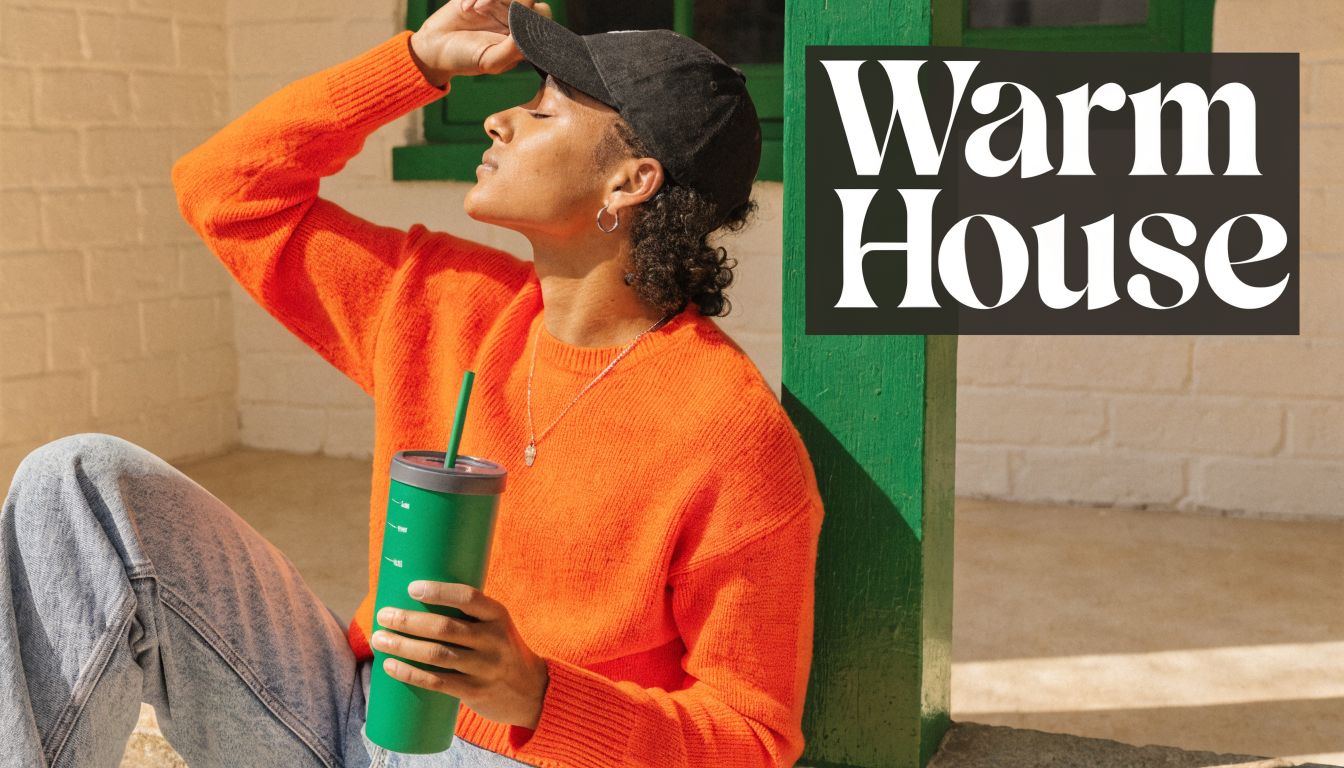 A person wearing a bright orange sweater and black cap holding a green tumbler while outdoors.